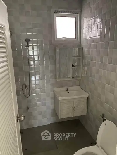 Compact bathroom with tiled walls, featuring a shower, sink, and toilet under a small window.