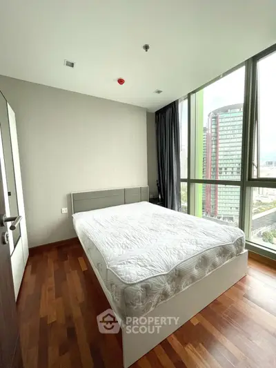 Modern bedroom with large windows and city view, featuring a sleek bed and wooden flooring.