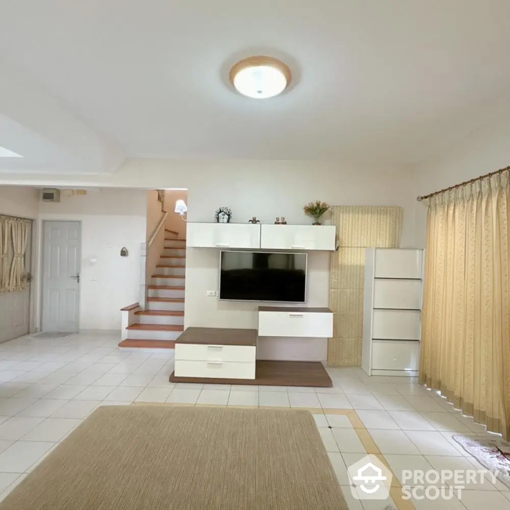 Spacious living room with modern entertainment unit, tiled flooring, and elegant staircase leading to upper level, bathed in natural light from large windows.