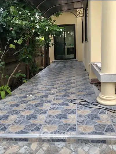Charming exterior walkway with tiled flooring and lush greenery leading to a modern entrance.