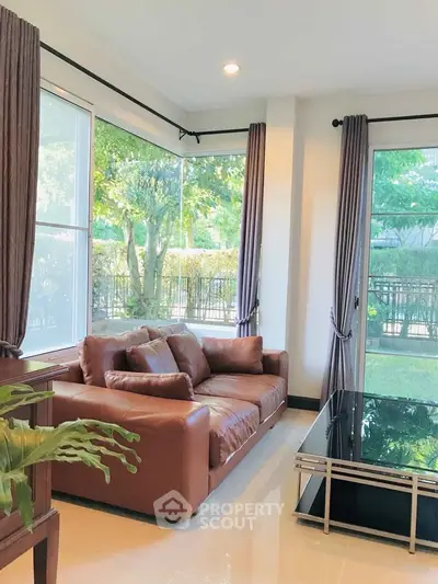 Bright living room with large windows and brown leather sofa overlooking lush garden.