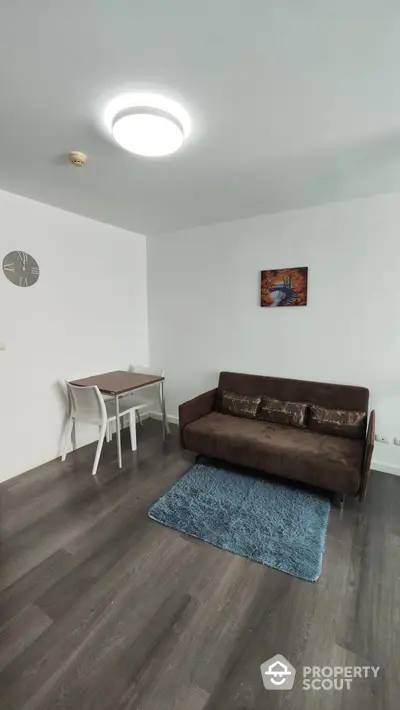 Modern minimalist living room with sleek dark wooden flooring, cozy brown sofa, vibrant blue rug, and a simple dining area, perfect for urban living.