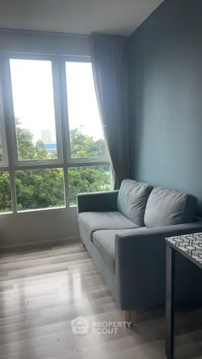 Cozy living room with large windows and a comfortable gray sofa, offering a serene view of lush greenery.