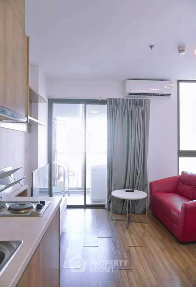 Modern apartment interior with compact kitchen and cozy living area featuring red sofa and balcony access.