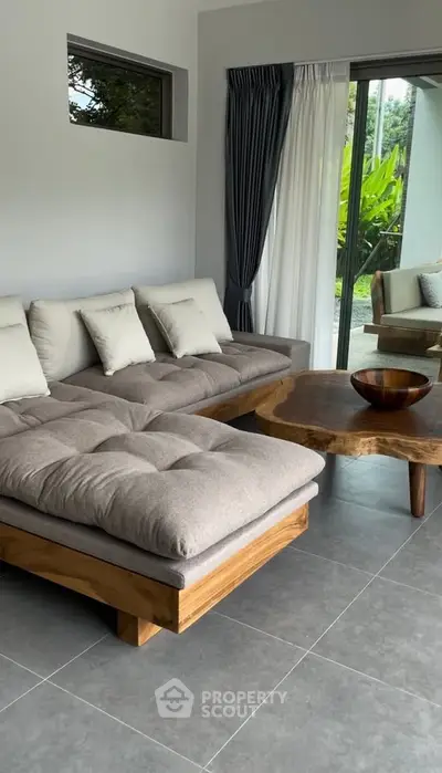 Modern living room with stylish gray sofa and wooden coffee table, overlooking lush garden through glass doors.