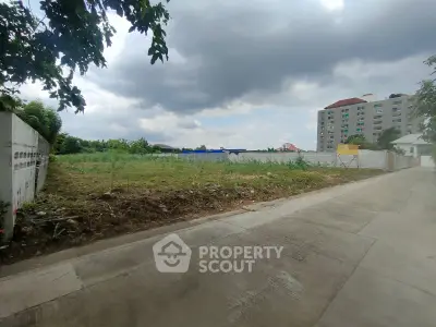 Vacant land plot with potential for development, adjacent to a residential building, under a cloudy sky.
