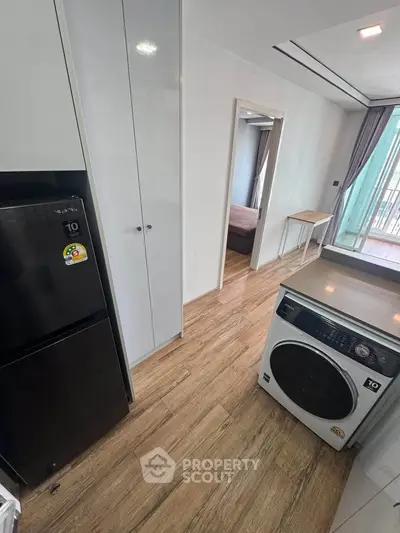 Modern apartment interior with washing machine and fridge, featuring wooden flooring and natural light.