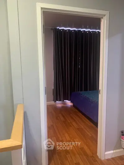 Cozy bedroom with wooden flooring and dark curtains, viewed through an open door.