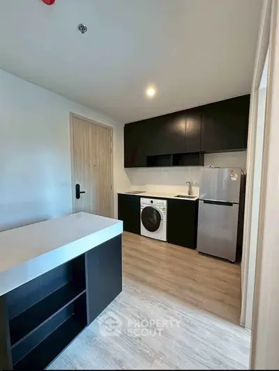 Modern kitchen with sleek black cabinets, washing machine, and compact fridge in a stylish apartment.