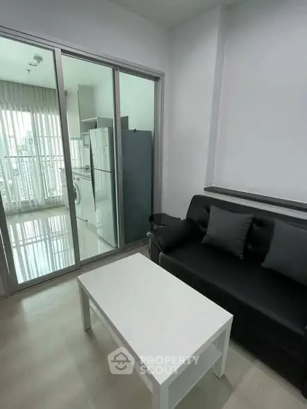 Modern living room with sleek black sofa and white coffee table, adjacent to a bright kitchen area.