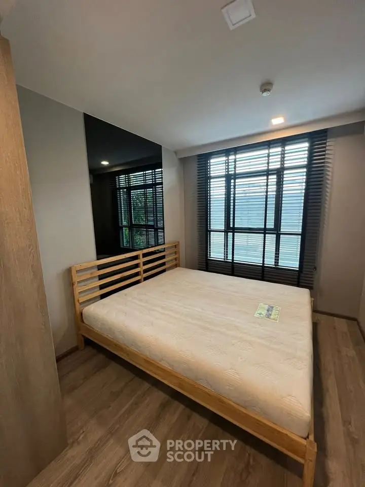 Modern bedroom with wooden flooring and large window, featuring a minimalist bed frame and natural light.