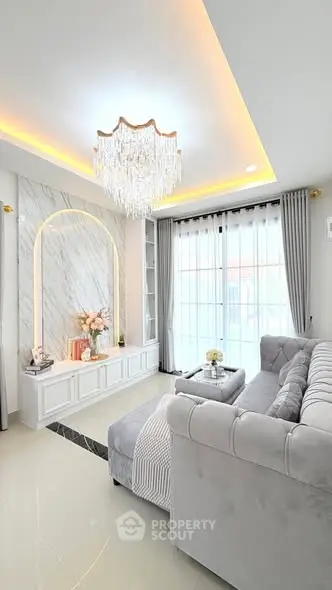 Luxurious living room with elegant chandelier and plush gray sofa, featuring modern decor and large windows.