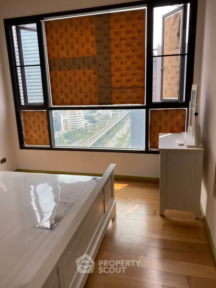 Bright bedroom with large windows offering city views, featuring wooden flooring and modern furniture.