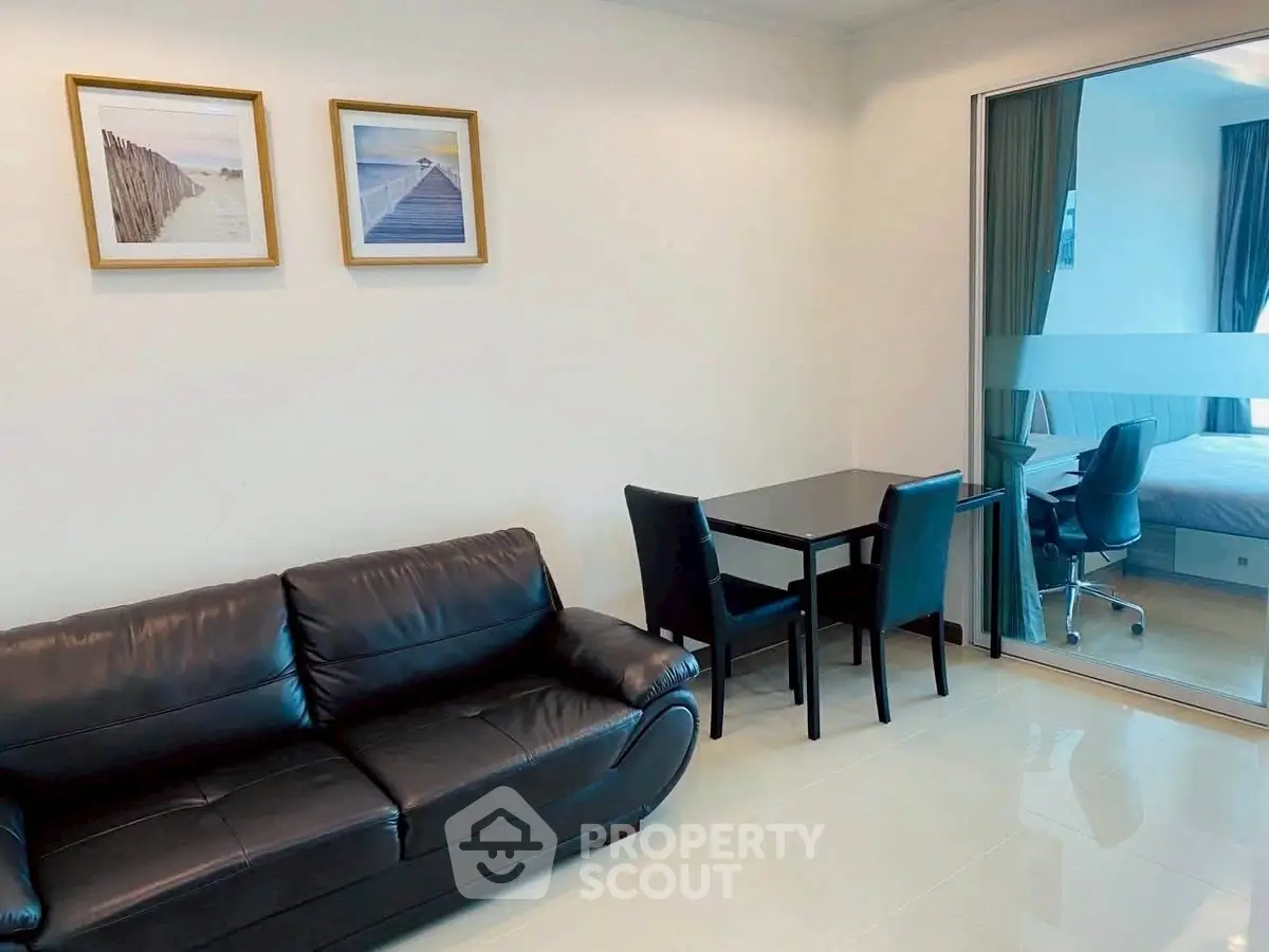 Modern living room with black leather sofa and glass partition leading to bedroom.