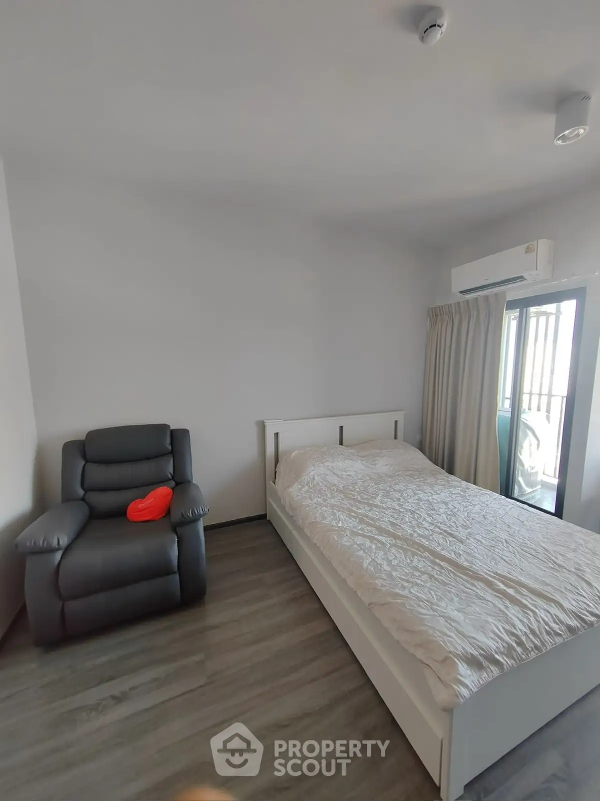 Modern bedroom with cozy bed and recliner, featuring natural light and sleek flooring.
