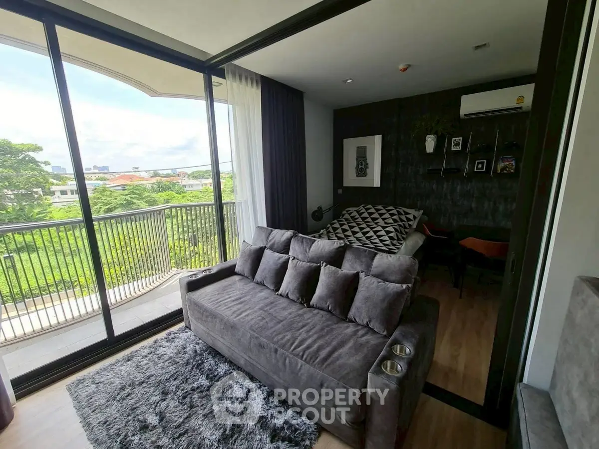 Modern living room with large windows and balcony overlooking lush greenery, featuring stylish decor and cozy seating.