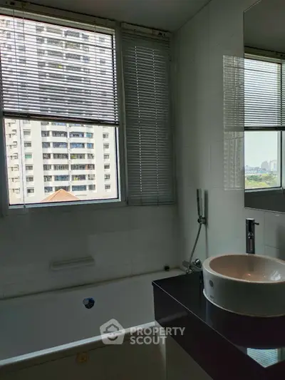 Modern bathroom with city view, featuring sleek sink and bathtub under large window.