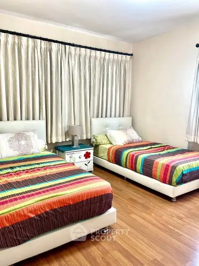 Bright bedroom with twin beds and colorful bedding, featuring wooden flooring and elegant curtains.