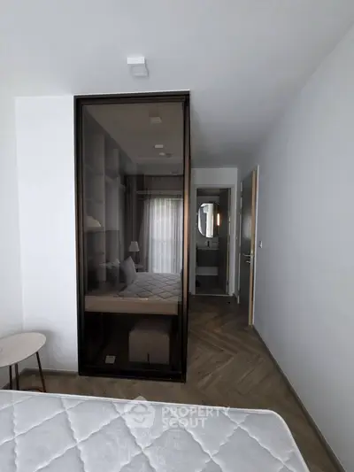 Modern bedroom with glass wardrobe and herringbone flooring, leading to a sleek bathroom.