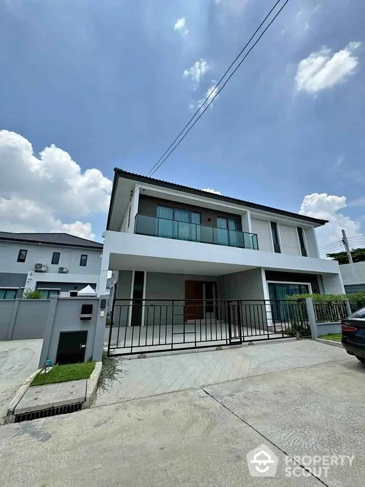 Modern two-story house with balcony and gated driveway under a clear blue sky.