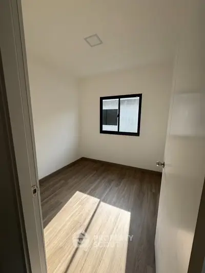 Minimalist bedroom with wooden flooring and natural light through window.