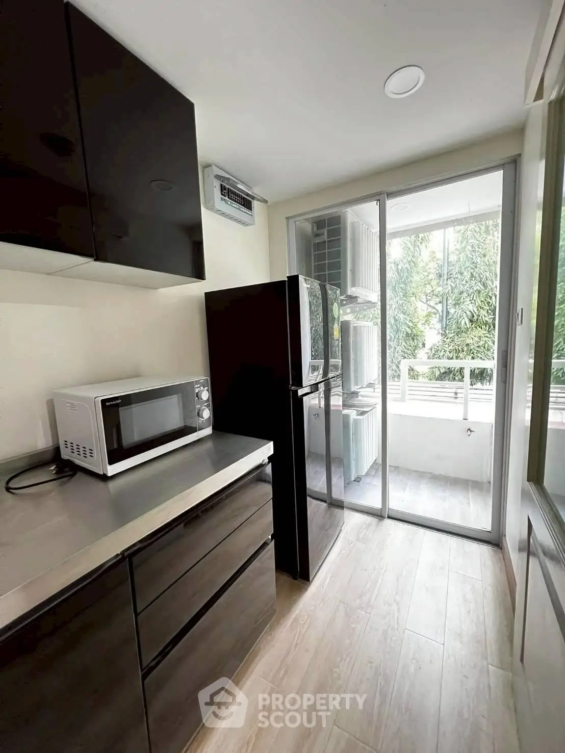 Modern kitchen with sleek cabinets, microwave, and fridge, leading to a bright balcony with greenery view.