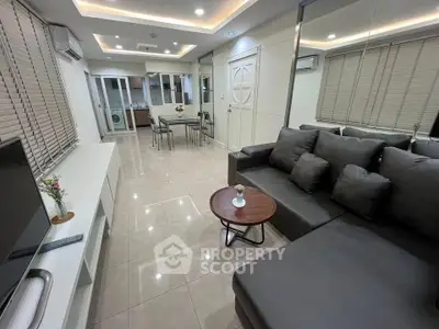 Modern living room with sleek gray sofa and open layout, featuring a dining area and washing machine in the background.