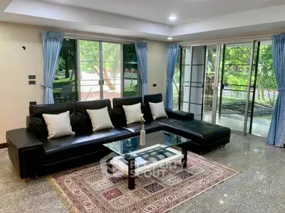 Spacious living room with modern black leather sofa and large windows overlooking lush greenery.