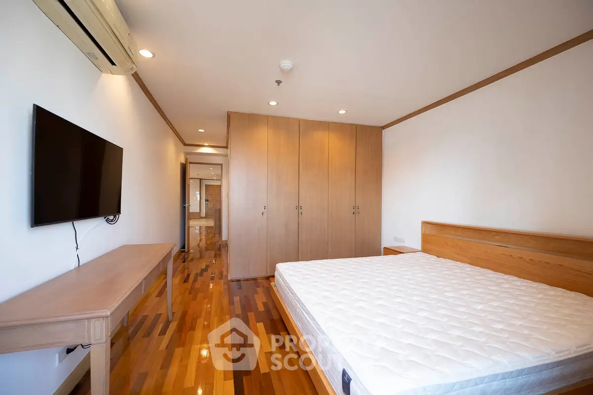 Spacious bedroom with wooden flooring, built-in wardrobe, and wall-mounted TV in modern apartment.
