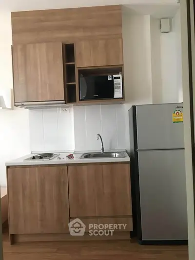 Modern compact kitchen with wooden cabinets, microwave, and fridge in a cozy apartment.