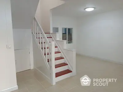 Spacious and bright living area with elegant red-accented staircase leading to the upper level, featuring pristine white walls and ample lighting.