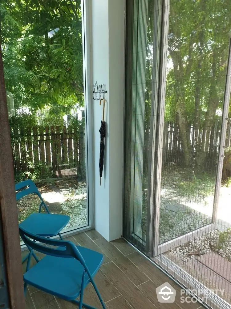 Charming balcony space with vibrant blue chairs overlooking a serene private garden, framed by floor-to-ceiling glass doors for an indoor-outdoor living experience.