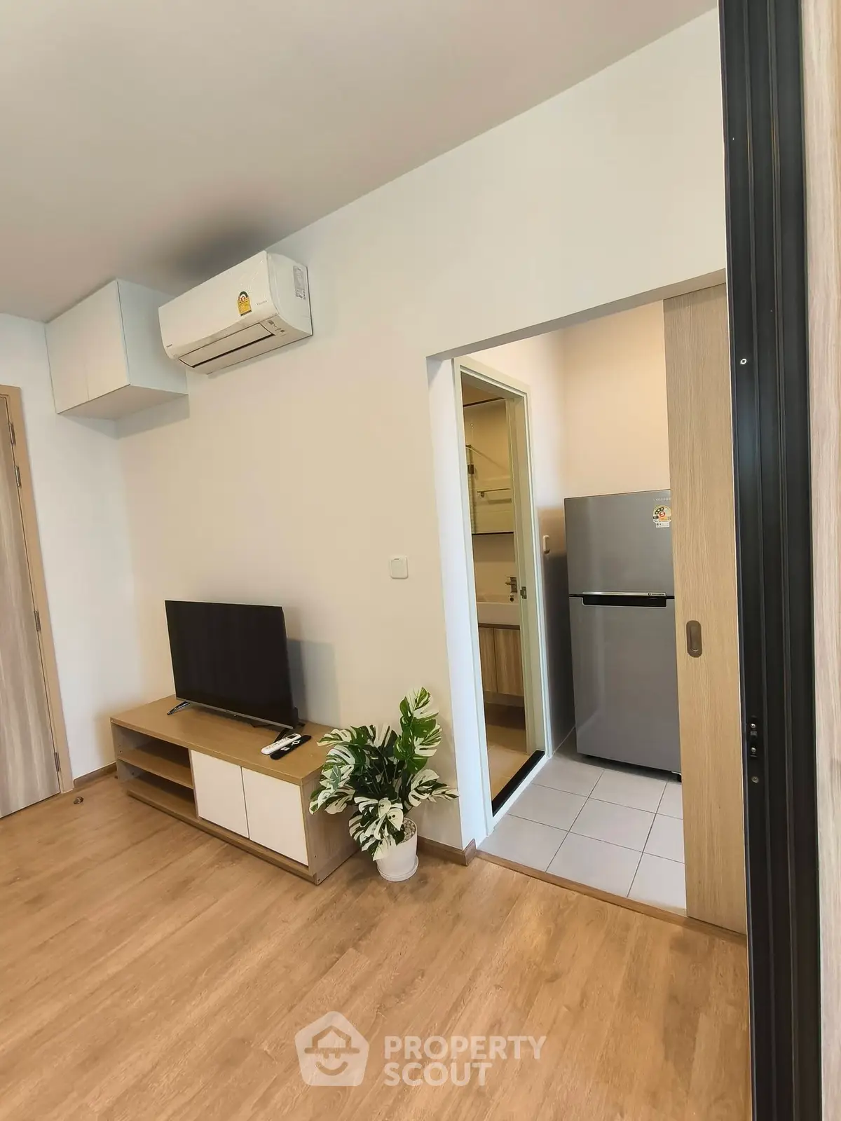 Modern living room with TV, air conditioning, and view into a compact kitchen with fridge.