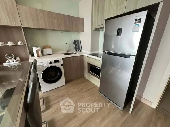 Modern kitchen with sleek appliances and wooden cabinetry, featuring a washing machine and stainless steel fridge.