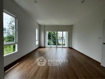 Spacious empty living room with large windows and wooden flooring, offering a serene view of greenery.