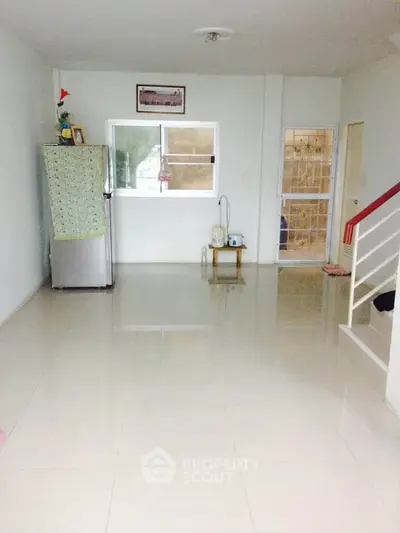 Spacious living room with tiled flooring and natural light, featuring a staircase and a covered refrigerator.