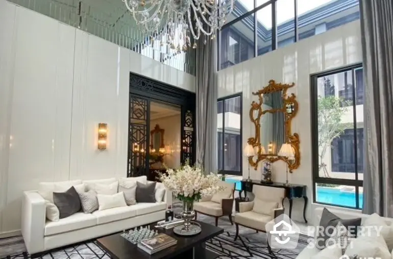 Luxurious living room with high ceilings, elegant chandelier, and opulent gold-framed mirror, leading to a serene pool view.