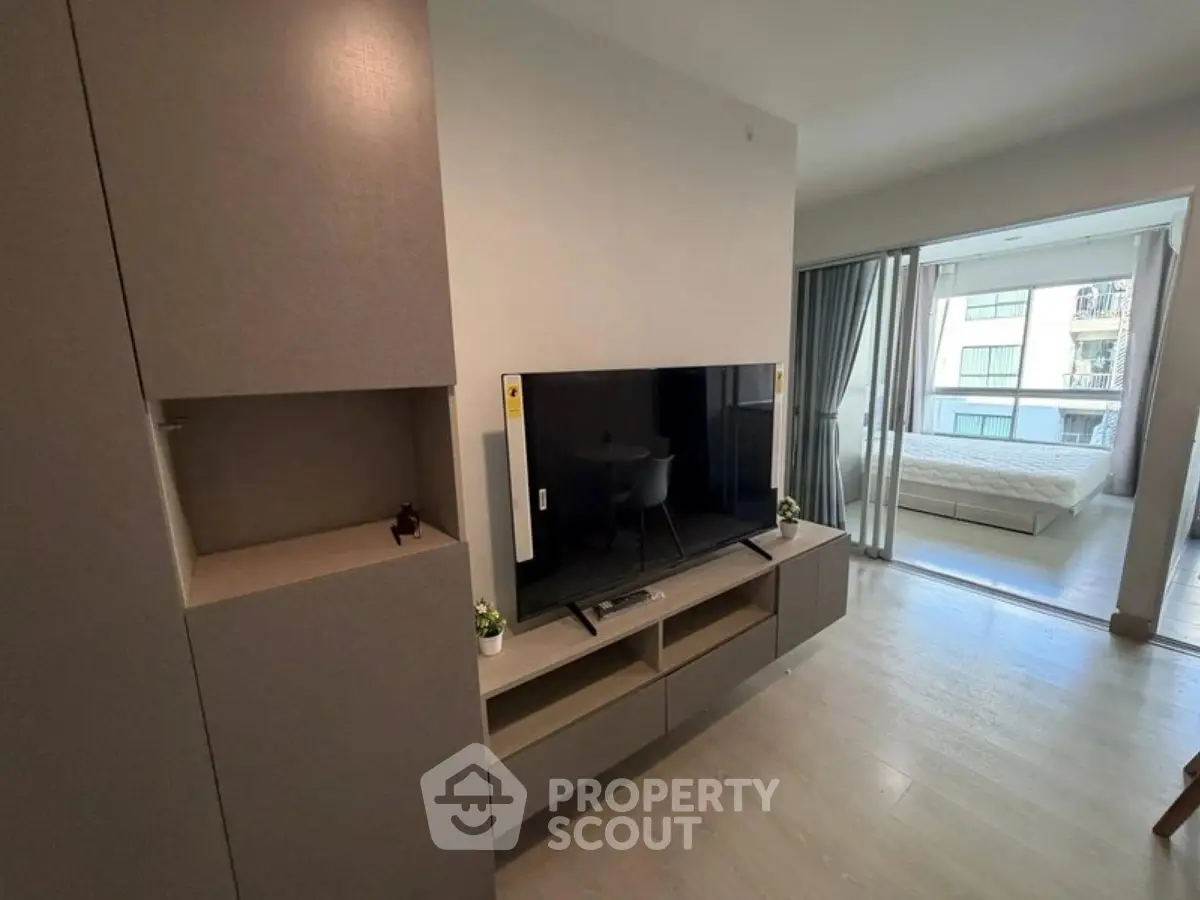 Modern living room with sleek TV unit and view into a bright bedroom with balcony access.