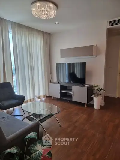 Modern living room with elegant chandelier and large TV, featuring stylish furniture and floor-to-ceiling windows.