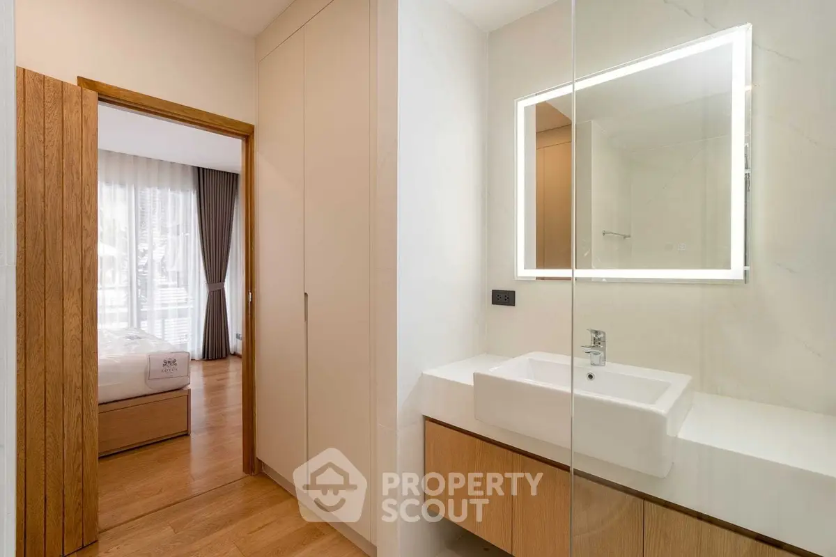 Modern bathroom with illuminated mirror and wooden accents, adjacent to a cozy bedroom with natural light.