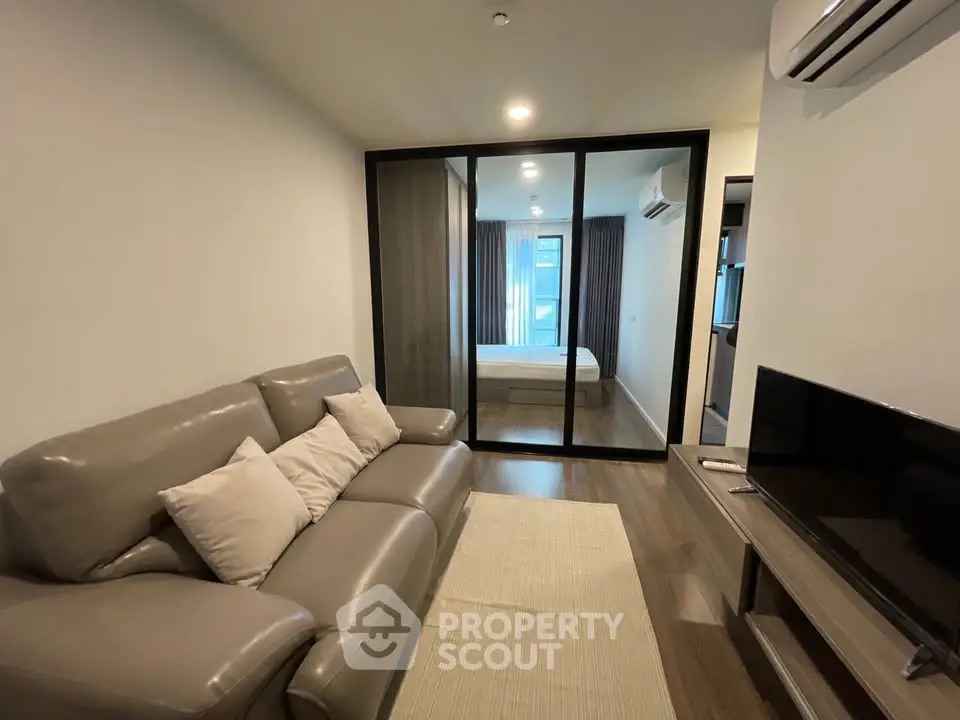 Modern living room with sleek glass partition leading to a cozy bedroom, featuring stylish furniture and air conditioning.