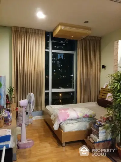 Cozy bedroom with wooden flooring, large window offering city views at night, and elegant drapery, perfect for urban living.