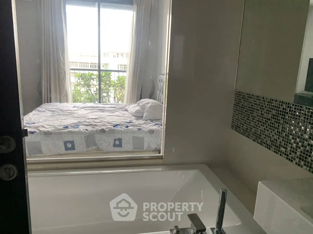 Modern bathroom with a view into a cozy bedroom, featuring a sleek bathtub and large window.