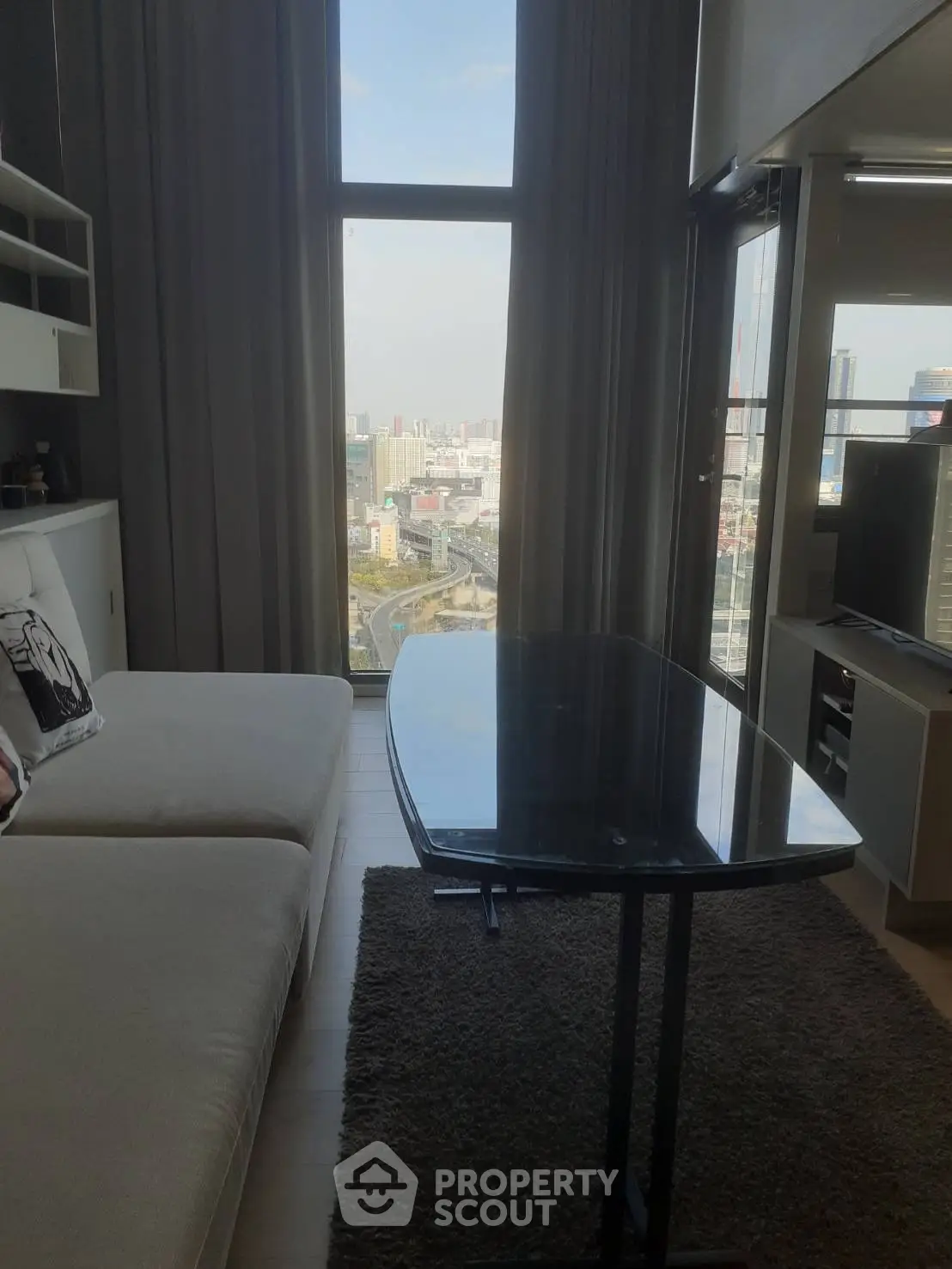 Modern living room with large window offering stunning city view, featuring cozy sofa and sleek glass table.