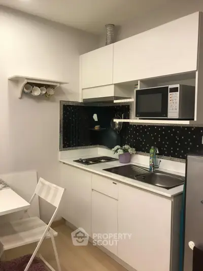 Modern compact kitchen with sleek white cabinets and black backsplash, featuring a microwave and dining area.