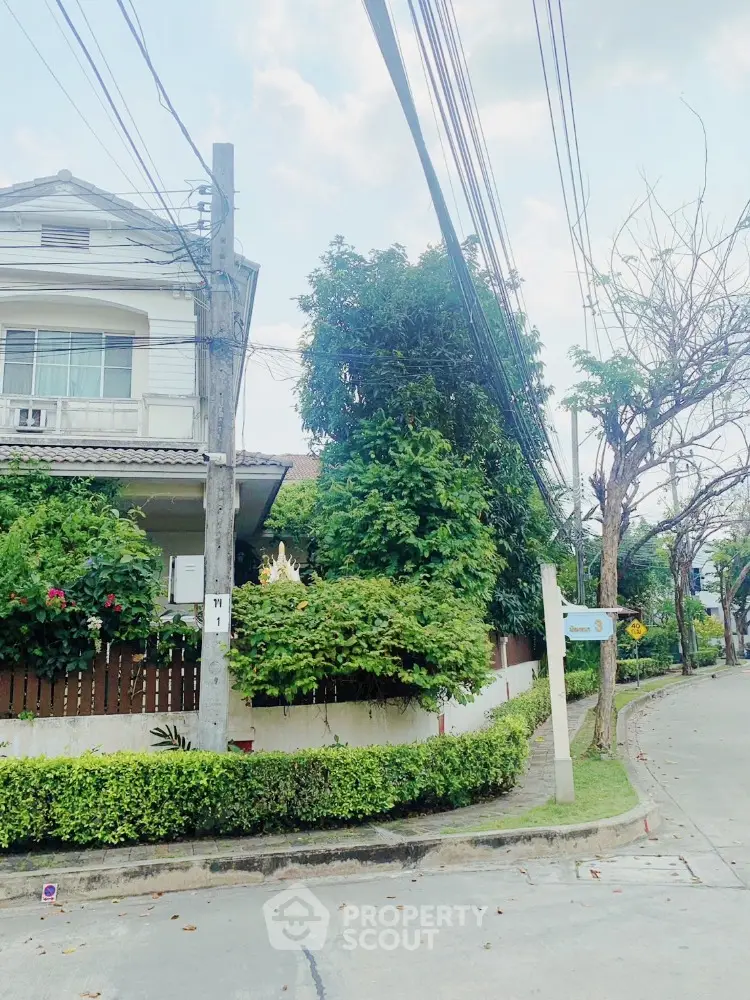 Charming two-story residence nestled in a lush, green neighborhood, boasting a well-manicured hedge and vibrant flora, complemented by a serene, tree-lined street.