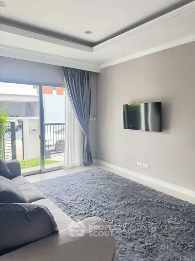 Modern living room with plush carpet, wall-mounted TV, and sliding glass doors leading to a balcony.