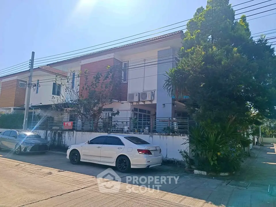 Modern townhouse with cars parked in front, featuring a lush tree and clear blue sky.