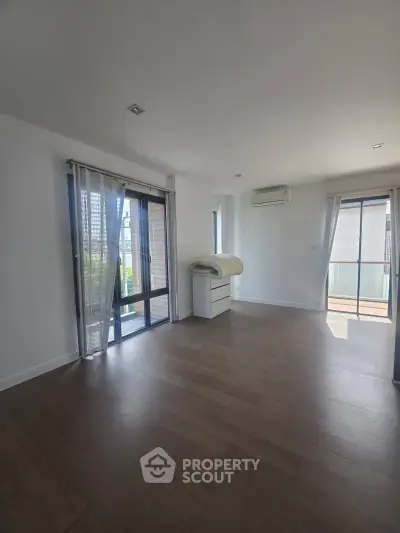 Spacious modern living room with large windows and balcony access, featuring sleek hardwood floors and abundant natural light.