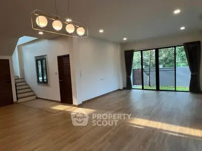 Spacious living room with modern lighting, large windows, and hardwood floors, leading to an inviting staircase and outdoor view.